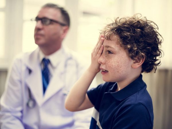 Little boy holding right hand over right eye for eye exam with doctor in the background