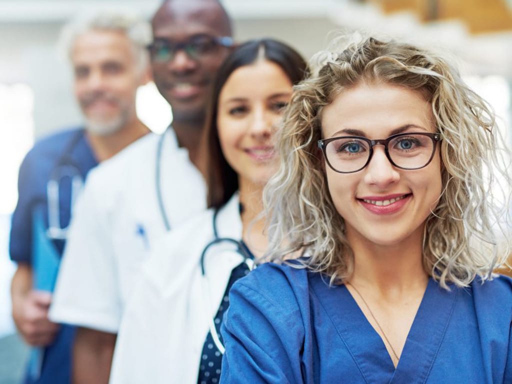 Curly haired blonde girl in glasses wearing blue scrubs with coworkers standing behind her