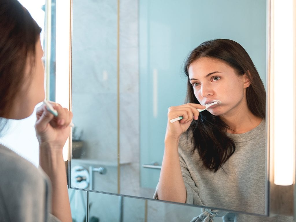 Dark haired girl in grey sweater looking in mirror brushing her teeth