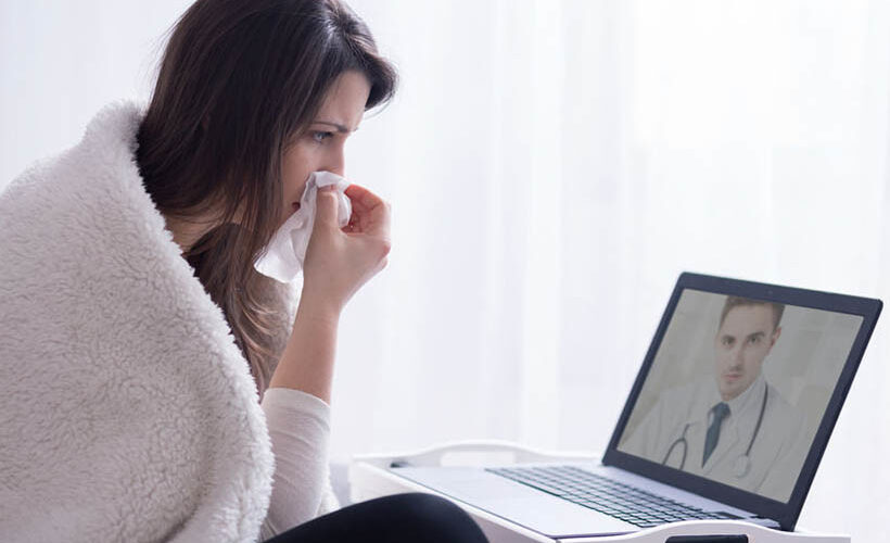 Sick woman wrapped in a blanket with a tissue at her nose and looking at a doctor through her computer screen