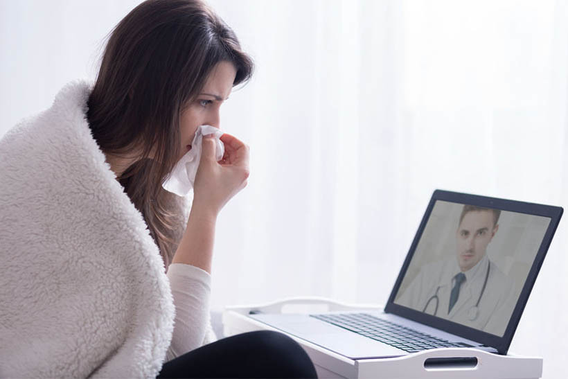 Sick woman wrapped in a blanket with a tissue at her nose and looking at a doctor through her computer screen