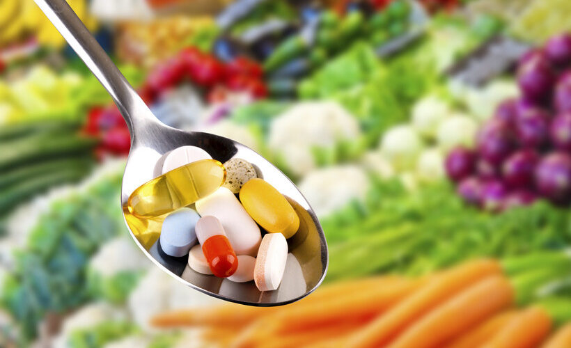vitamins in a silver spoon in the forefront with fruits and vegetables in the background