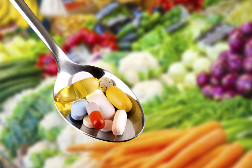vitamins in a silver spoon in the forefront with fruits and vegetables in the background