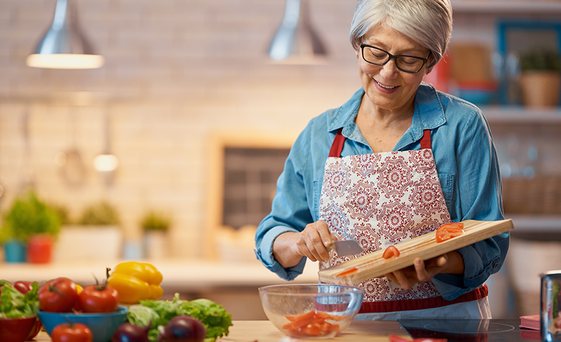 Older woman standing in the kitchen prepping vegetables with a knife, cutting board and bowl