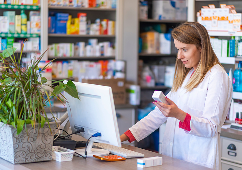 female pharmacist in lab coat holding a prescription box and looking at the computer