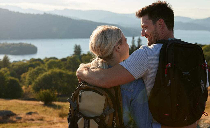 Dark haired man and blonde woman overlooking a lake in nature, wearing backpacks