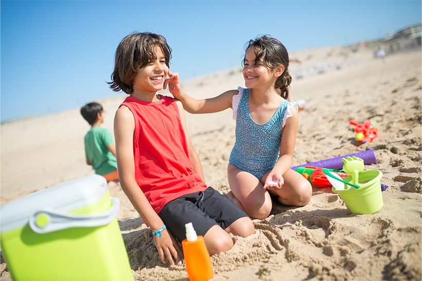 Two young kids on the beach with a girl putting sunscreen on a boy's face for protection.