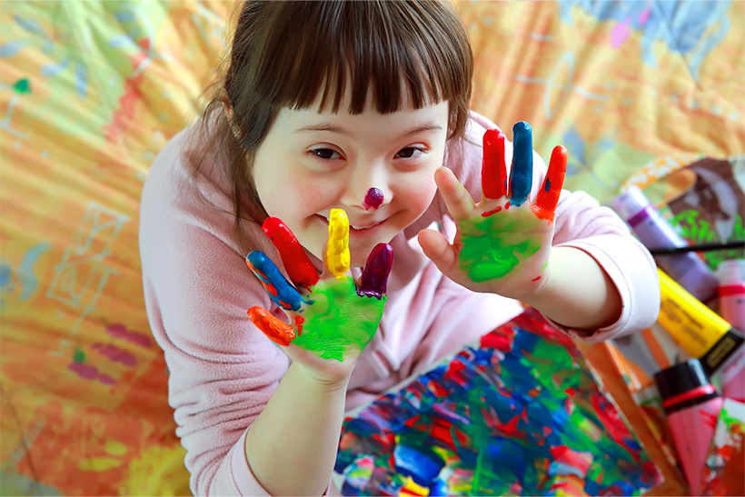 Girl with dark hair and Down Syndrome showing her hands to the camera that are covered with colorful paint while she is smiling